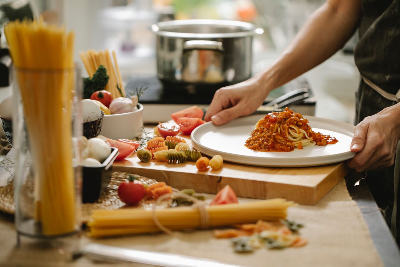 journey Crop anonymous cook standing at table with various ingredients and cooking pasta with meat and tomatoes